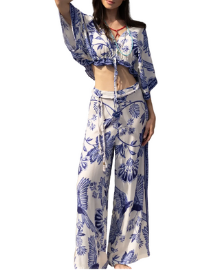 A woman models a matching two-piece bohemian-style set featuring a blue and white floral print. The set includes a cropped tie-front top with wide, draped sleeves and matching wide-leg pants, styled outdoors against a weathered wooden wall.