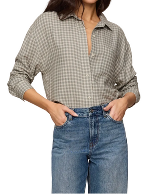 A woman wearing a muted sage green and gray gingham button-up shirt tucked into blue jeans, standing against a plain light beige background.