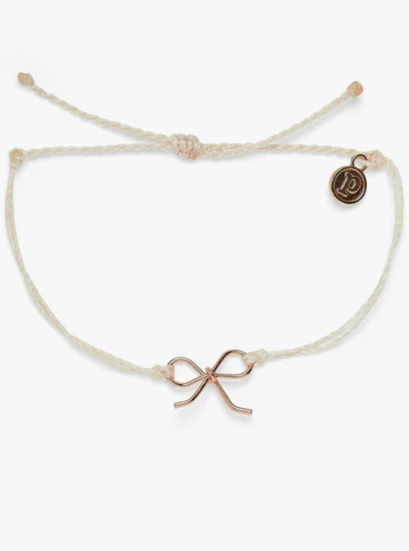 Bow Charm Rose Gold Bracelet in Vanilla  We’re knot kidding when we say our Bow Charm Bracelet is the most adorable style ever! This braid design features a delicate bow charm for the perfect piece to complete a cutesy-cool stack.  Wax-coated bracelet Silver: brass base with rhodium plating Rose Gold: brass base with rose gold plating Bow charm: 13mm (H) x 16mm (W) Adjustable from approximately 2-5 inches in diameter